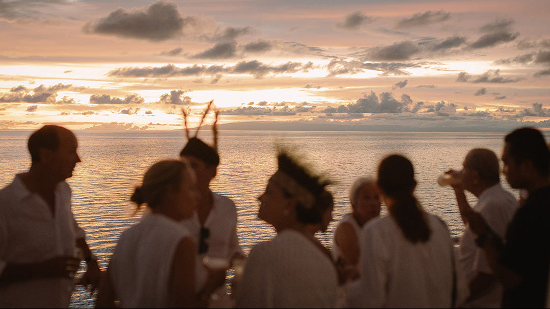 A group of people standing on a boat or deck at sunset, facing the ocean with a cloudy sky in the background. The scene is lit by warm golden hour light, but the people are somewhat silhouetted and out of focus.