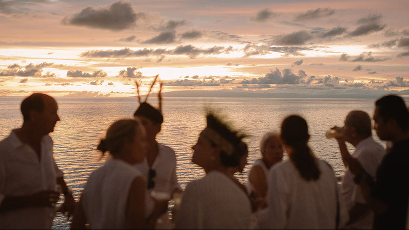 A group of people standing on a boat or deck at sunset, facing the ocean with a cloudy sky in the background. The scene is lit by warm golden hour light, but the people are somewhat silhouetted and out of focus.